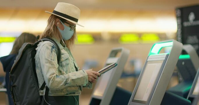Woman in airport. Female traveler in preventive face mask typing on touch screen of the self check-in station in empty international terminal. Flight boarding registration, COVID-19 pandemic, 4K USA