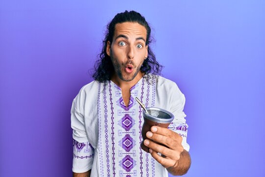 Young handsome man with long hair drinking mate infusion scared and amazed with open mouth for surprise, disbelief face