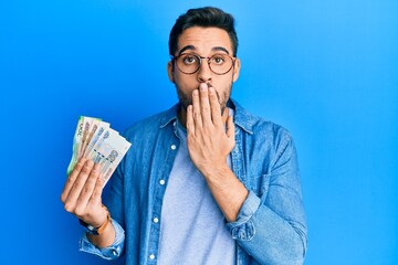 Young hispanic man holding russian ruble banknotes covering mouth with hand, shocked and afraid for...