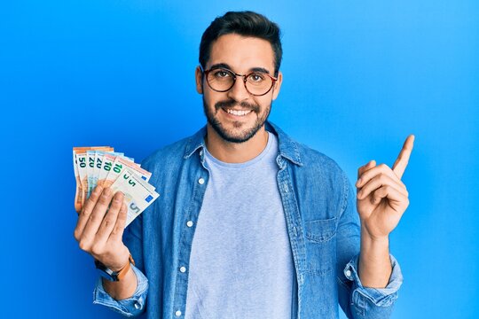 Young hispanic man holding euro banknotes smiling happy pointing with hand and finger to the side