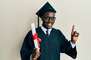 Handsome black man wearing graduation cap and ceremony robe holding diploma surprised with an idea...