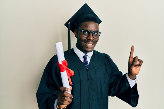 Handsome black man wearing graduation cap and ceremony robe holding diploma surprised with an idea or question pointing finger with happy face, number one - Powered by Adobe