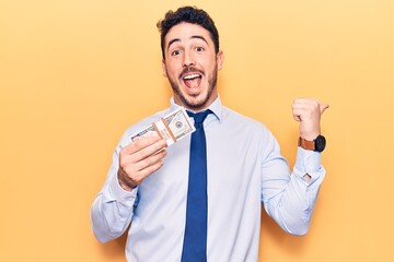 Young hispanic man wearing business clothes holding dollars pointing thumb up to the side smiling happy with open mouth