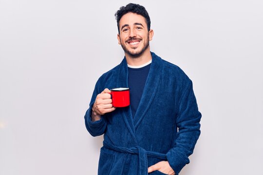 Young hispanic man wearing robe holding coffee looking positive and happy standing and smiling with a confident smile showing teeth