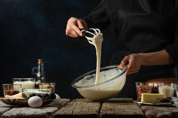Pastry chef kneads a dough with a whisk. Backstage of cooking waffle on rustic wooden table with ingredients on dark blue background. Frozen motion. Handmade dessert. Cooking process.