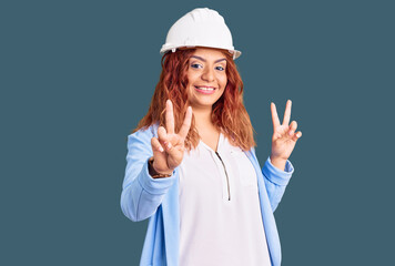 Young latin woman wearing architect hardhat smiling looking to the camera showing fingers doing victory sign. number two.