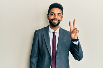 Handsome hispanic man with beard wearing business suit and tie smiling with happy face winking at the camera doing victory sign with fingers. number two.