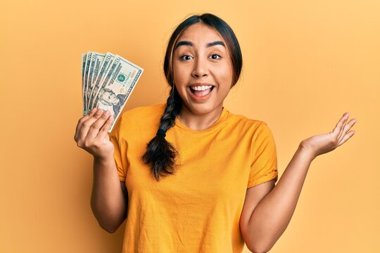Young latin woman holding dollars celebrating achievement with happy smile and winner expression with raised hand