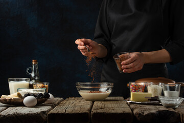 Fototapeta na wymiar Pastry chef pours cocoa into glass bowl. Backstage of cooking waffle on rustic wooden table with ingredients on dark blue background. Frozen motion. Handmade dessert. Cooking process.