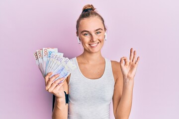 Beautiful blonde sport woman holding 20 swedish krona banknotes doing ok sign with fingers, smiling friendly gesturing excellent symbol