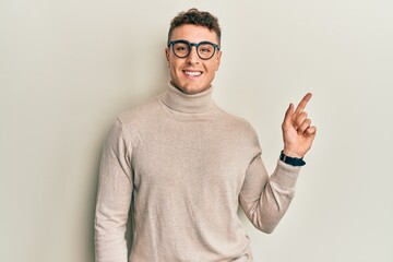 Fototapeta premium Hispanic young man wearing casual turtleneck sweater with a big smile on face, pointing with hand finger to the side looking at the camera.