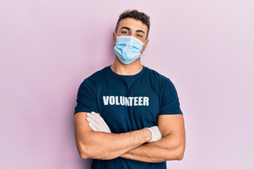 Hispanic young man wearing medical mask and volunteer t shirt happy face smiling with crossed arms looking at the camera. positive person.