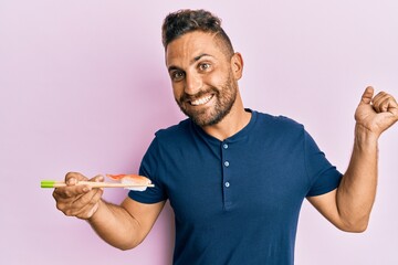 Handsome man with beard eating sushi using chopsticks screaming proud, celebrating victory and success very excited with raised arm