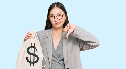 Young chinese woman wearing business suit holding dollars bag with angry face, negative sign...