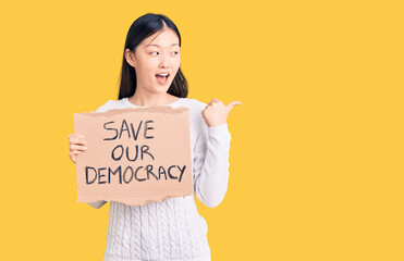 Young beautiful chinese woman holding save our democracy cardboard banner pointing thumb up to the...