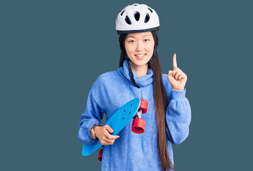 Young beautiful chinese woman wearing security helmet holding skate surprised with an idea or question pointing finger with happy face, number one
