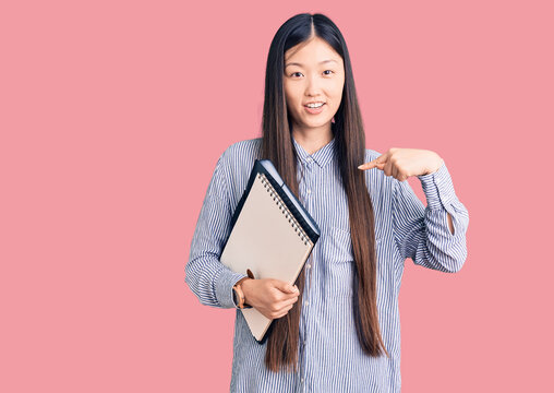 Young beautiful chinese woman holding notebook pointing finger to one self smiling happy and proud