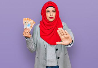 Beautiful hispanic woman wearing islamic hijab holding 10 swiss franc banknotes with open hand doing stop sign with serious and confident expression, defense gesture