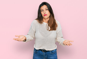 Beautiful hispanic woman wearing casual sweater clueless and confused expression with arms and hands raised. doubt concept.