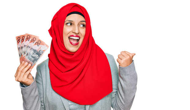 Beautiful hispanic woman wearing islamic hijab holding australian dollars banknotes pointing thumb up to the side smiling happy with open mouth