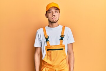 Hispanic young man wearing handyman uniform puffing cheeks with funny face. mouth inflated with air, crazy expression.