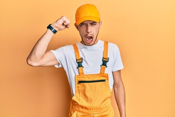 Hispanic young man wearing handyman uniform angry and mad raising fist frustrated and furious while shouting with anger. rage and aggressive concept.