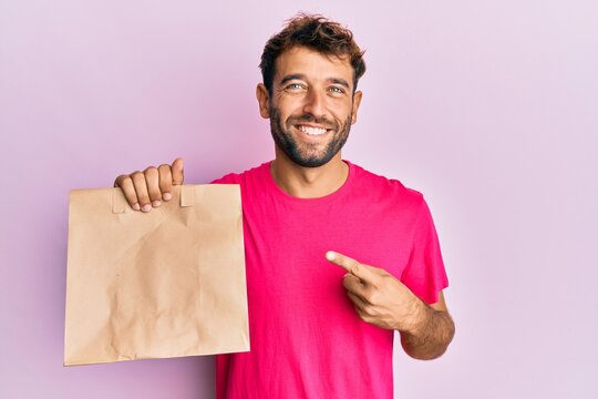 Handsome man with beard holding take away paper bag smiling happy pointing with hand and finger