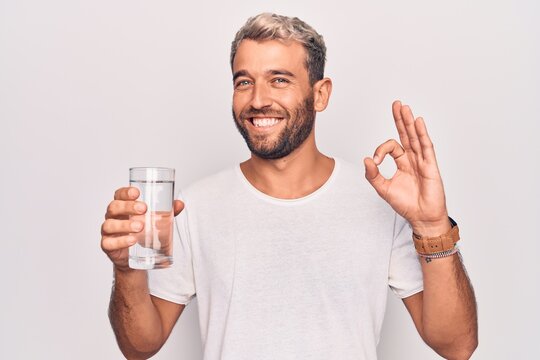 Handsome blond man with beard drinking glass of water to refreshment over white background doing ok sign with fingers, smiling friendly gesturing excellent symbol