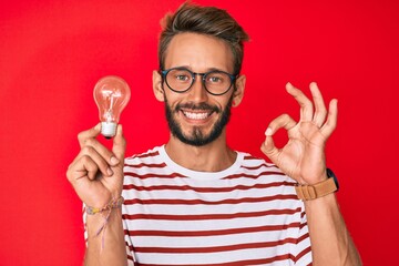 Handsome caucasian man with beard holding lightbulb for inspiration and idea doing ok sign with fingers, smiling friendly gesturing excellent symbol