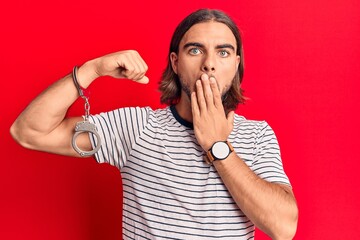 Young handsome man wearing prisoner handcuffs covering mouth with hand, shocked and afraid for...