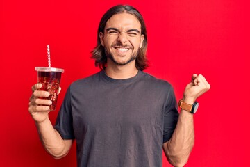 Young handsome man drinking glass of cola beverage screaming proud, celebrating victory and success...