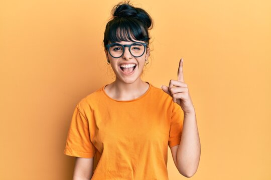 Young hispanic girl wearing casual clothes and glasses pointing finger up with successful idea. exited and happy. number one.