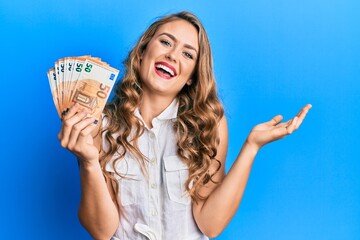 Young blonde girl holding euro banknotes celebrating achievement with happy smile and winner expression with raised hand - obrazy, fototapety, plakaty