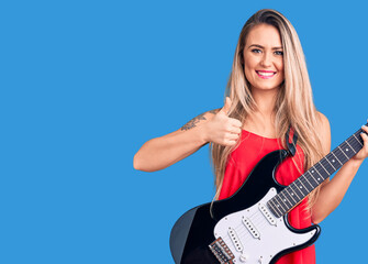 Fototapeta premium Young beautiful blonde woman playing electric guitar smiling happy and positive, thumb up doing excellent and approval sign