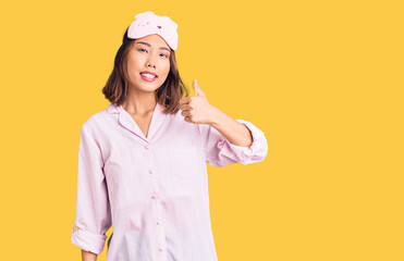 Young beautiful chinese girl wearing sleep mask and pajama doing happy thumbs up gesture with hand. approving expression looking at the camera showing success.