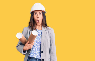 Young beautiful brunette woman wearing architect hardhat holding blueprint scared and amazed with open mouth for surprise, disbelief face