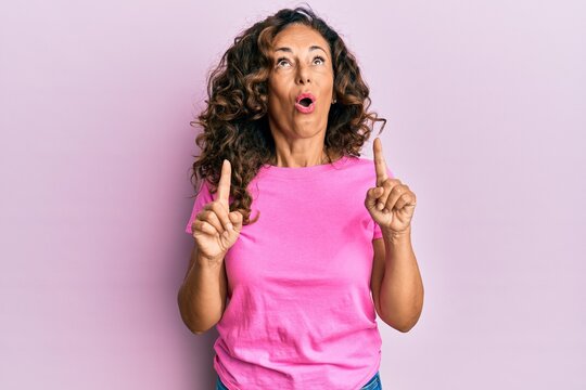 Middle age hispanic woman wearing casual clothes amazed and surprised looking up and pointing with fingers and raised arms.