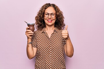 Middle age beautiful woman drinking cup of mate over isolated pink background smiling happy and positive, thumb up doing excellent and approval sign