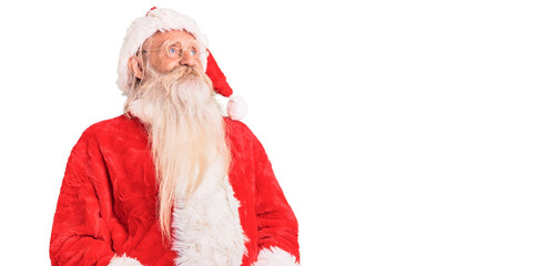 Old senior man with grey hair and long beard wearing traditional santa claus costume looking away to side with smile on face, natural expression. laughing confident.