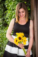 Caucasian Armenian young woman holding sunflowers wearing a black and white dress standing next to a wall of ivy