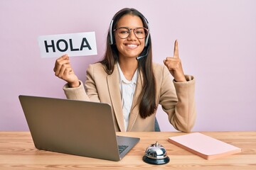 Beautiful hispanic woman wearing operator headset holding hola greenting smiling with an idea or question pointing finger with happy face, number one © Krakenimages.com