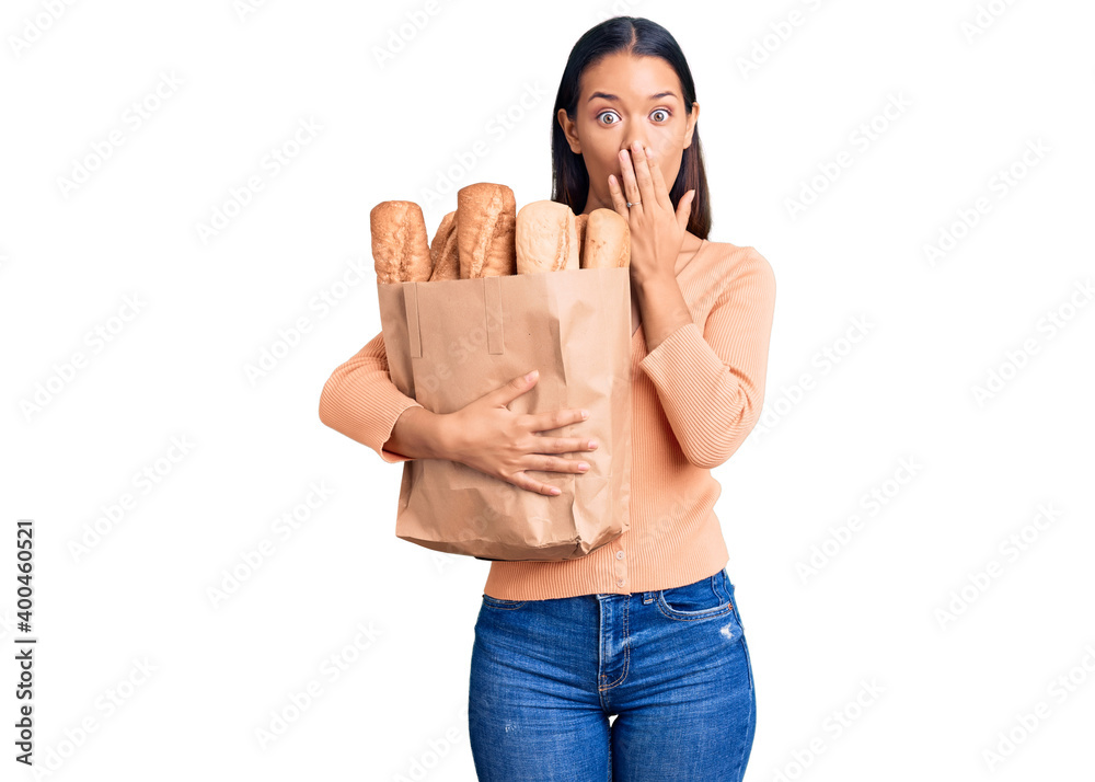 Sticker Young beautiful latin girl holding paper bag with bread covering mouth with hand, shocked and afraid for mistake. surprised expression