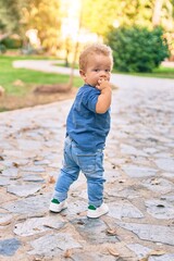 Sad little boy putting fingers on mouth touching gums because toothache at the park on a sunny day. Beautiful blonde hair male toddler in pain for new baby teeth outdoors