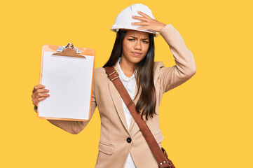 Beautiful hispanic woman architect showing black space on clipboard stressed and frustrated with hand on head, surprised and angry face