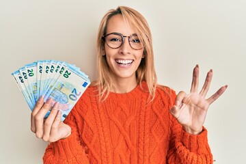 Beautiful blonde woman holding 20 euro banknotes doing ok sign with fingers, smiling friendly...