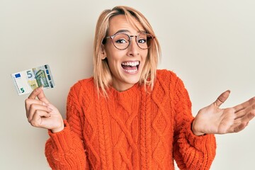 Beautiful blonde woman holding 5 euro banknote celebrating achievement with happy smile and winner...