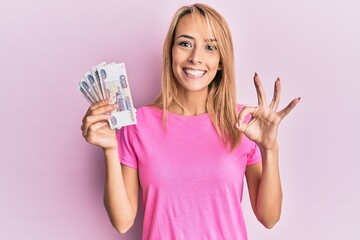 Beautiful blonde woman holding 500 russian ruble banknotes doing ok sign with fingers, smiling friendly gesturing excellent symbol