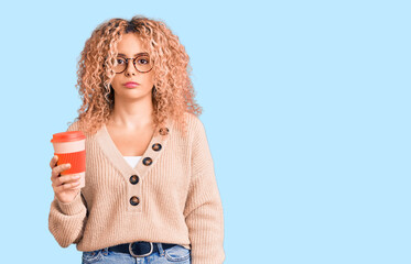 Young blonde woman with curly hair wearing glasses and drinking a cup of coffee thinking attitude and sober expression looking self confident