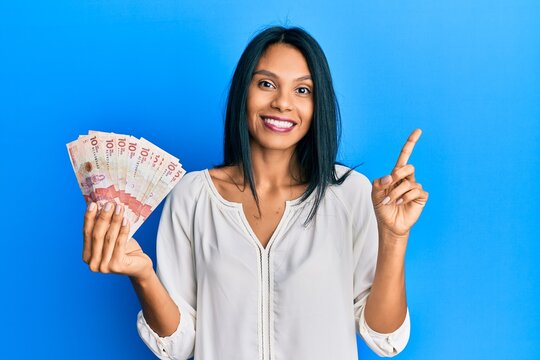 Young african american woman holding 10 colombian pesos banknotes cheerful with a smile on face pointing with hand and finger up to the side with happy and natural expression