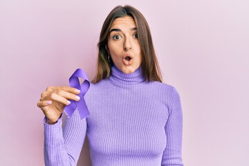 Young hispanic woman holding purple ribbon awareness scared and amazed with open mouth for...